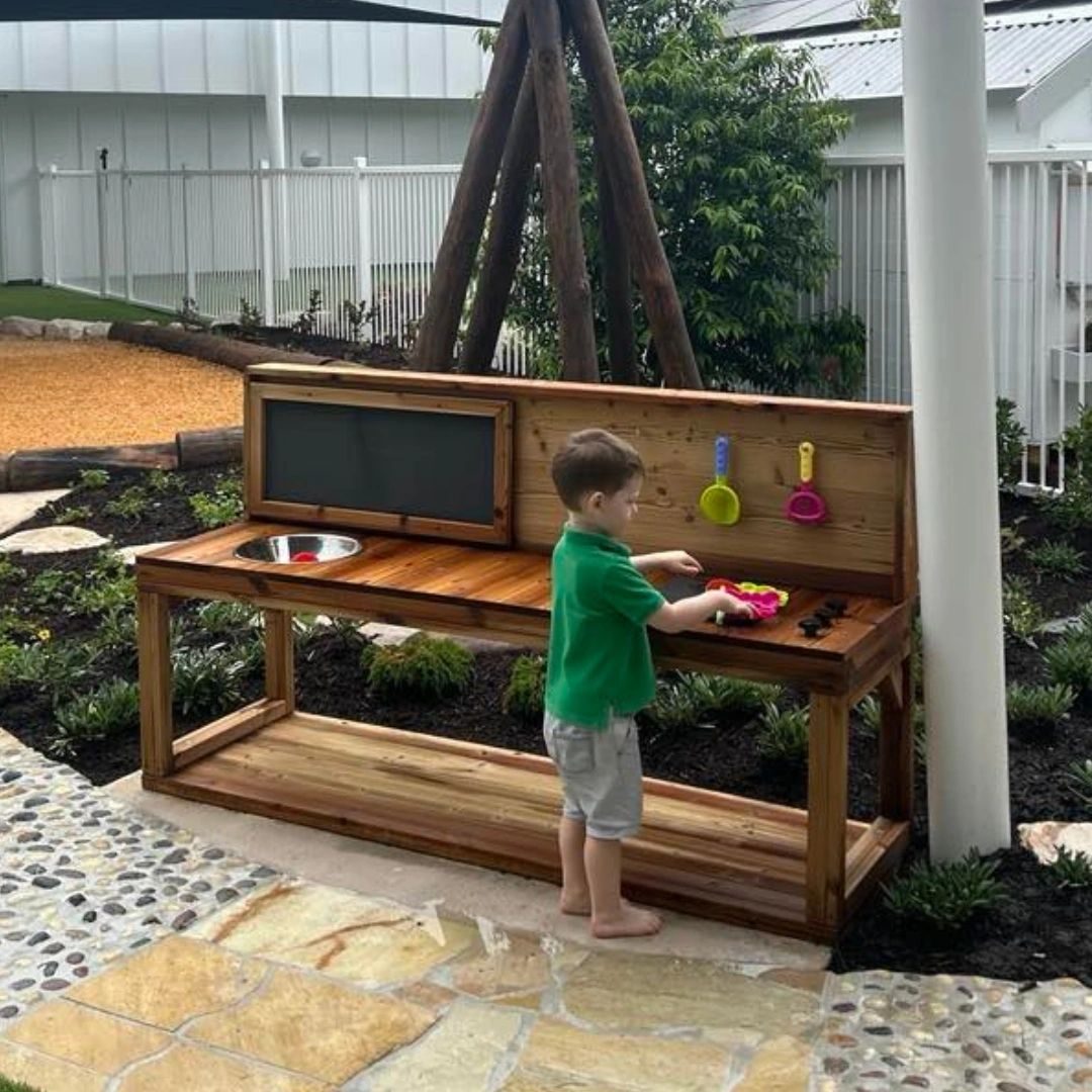 Child playing at outdoor timber mud kitchen with chalkboard and utensils